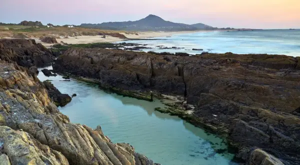 Aguas cristalinas en la **playa das Furnas**, entre rocas volcánicas y arena dorada, con el mar al fondo y una montaña.