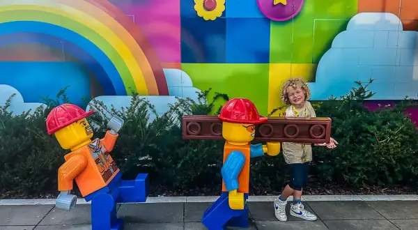 Chłopiec z uśmiechem pozuje z dwoma wielkimi ludzikami LEGO na tle kolorowego muralu. To świetne billund atrakcje dla całej rodziny!