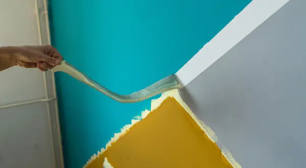 Hand entfernt Klebeband, um eine scharfe Kante beim Farbe streichen zu erzielen. Türkis trifft auf Grau und Ockergelb.