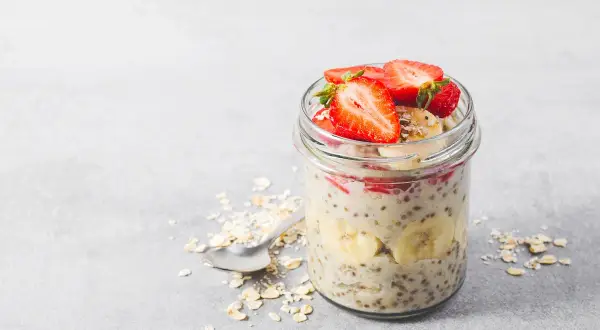 En burk med overnight oats recept toppad med jordgubbar och banan. Havregryn och en sked ligger bredvid.