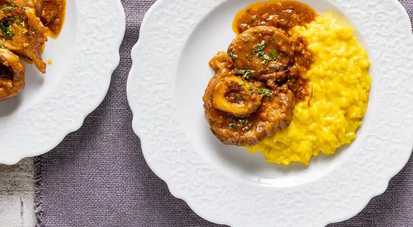 Ossobuco ricetta originale servito su un piatto bianco con risotto giallo allo zafferano.
