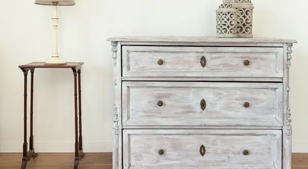 Un meuble cérusé patiné, avec ses tiroirs et ses ornements délicats, se dresse à côté d'une petite table d'appoint surmontée d'une lampe.