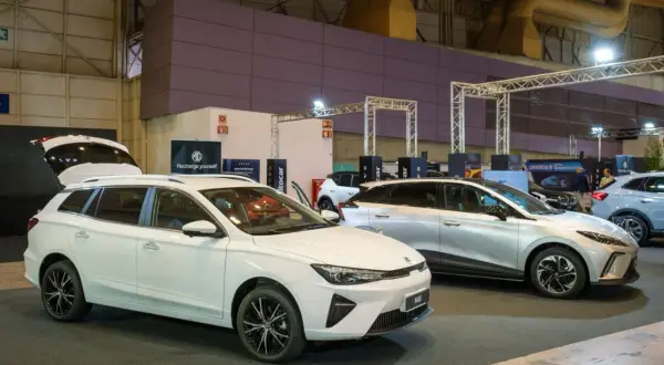 Białe kombi MG5 z otwartą klapą bagażnika i srebrny SUV MG na wystawie samochodowej.