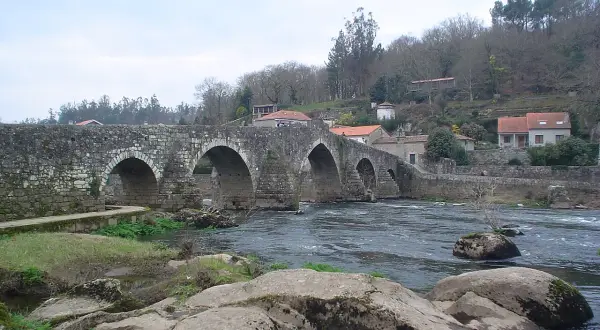 El ponte Maceira, un puente de piedra con arcos, cruza un río caudaloso. Casas con tejados rojos se asoman en la ladera.