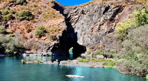 Un río de aguas turquesas fluye hacia un monte furado, una cueva natural en la roca, con vegetación y personas disfrutando del paisaje.