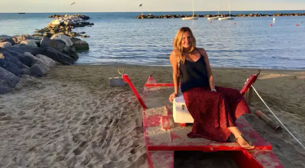Donna sorridente su una barca rossa sulla spiaggia di Cattolica, Emilia Romagna, con il mare e i faraglioni sullo sfondo.