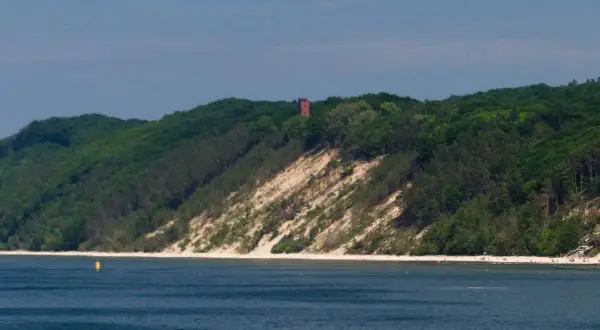 Zielone wzgórza porośnięte lasem schodzą ku morzu. Na szczycie wzniesienia, na tle błękitnego nieba, stoi ceglana wieża. Pomiędzy lasem a wodą rozciąga się piaszczysta plaża, tworząc białą górę.