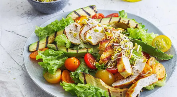 Gegrillter Hähnchensalat mit Avocado, Tomaten und Sprossen auf einem Teller.
