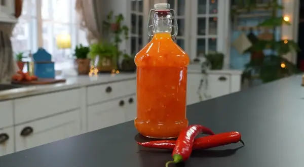 En flaska med hemmagjord sweet chili sås står på en mörk bänk, med två röda chilifrukter framför.