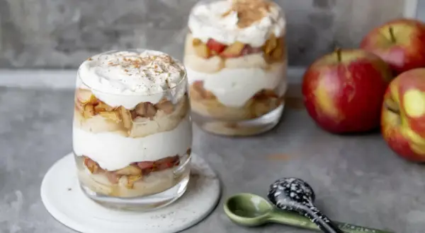 Délicieux tiramisu pomme biscuit cuillère, superposé dans des verres avec de la crème fouettée et de la cannelle. Des pommes rouges sont à côté.