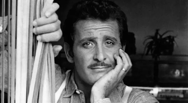 Domenico Modugno, con la mano appoggiata al viso, guarda pensieroso, mentre l'altra mano stringe delle corde.