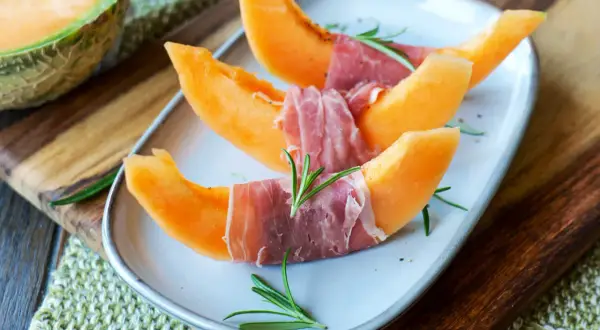 Melone mit Schinken, garniert mit Rosmarin, auf einem Teller. Ein Klassiker, der süße und salzige Aromen vereint.