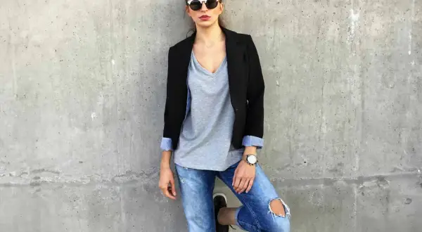Stylisches Outfit mit Blazer: Frau in Jeans, grauem V-Shirt und Sonnenbrille lehnt an Betonwand.