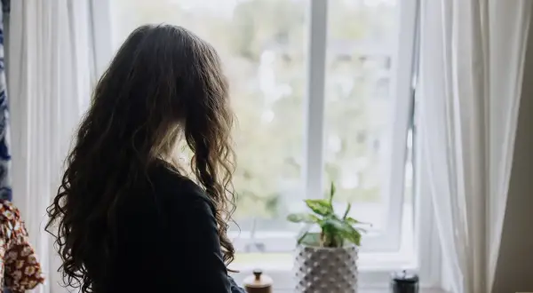 Flicka med långt hår tittar ut genom fönstret. En planta står på fönsterbrädan. Bilden kan symbolisera emotionell utveckling 3 år.