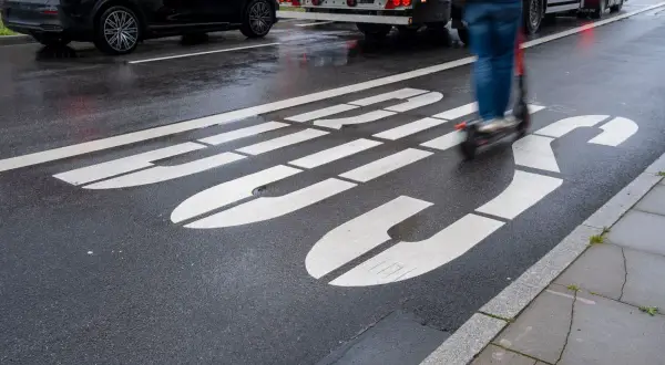 Jazda hulajnogą po buspasie grozi mandatem. Na mokrej drodze widać białe linie wyznaczające pas oraz poruszającego się po nim użytkownika hulajnogi.