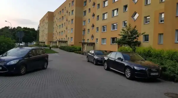 Na drodze osiedlowej zaparkowane są samochody przed blokiem z żółtą elewacją i nietypowymi oknami.