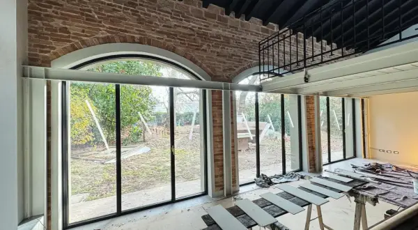 Interno in fase di ristrutturazione con ampie vetrate sul giardino. In primo piano, assi di legno appoggiate su cavalletti, segno di lavori di miglioramento della casa in corso.