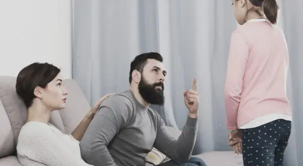 Una bambina ascolta il padre che la ammonisce, mentre la madre, con un'espressione preoccupata, le appoggia una mano sulla spalla, forse per alleviare la sua paura di perdere i genitori.