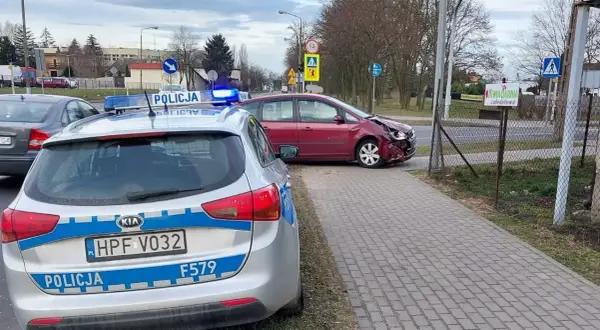 Samochód policyjny z włączonymi sygnałami świetlnymi przy drodze, gdzie doszło do kolizji drogowej. Czerwony samochód ma uszkodzony przód.