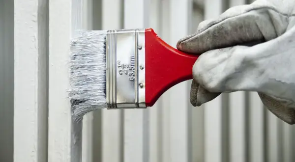 Mano guantata applica vernici industriali bianche su una ringhiera metallica con un pennello.