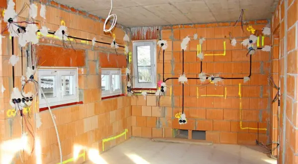 W trakcie budowy domu, widoczne są **domowe instalacje elektryczne** na ścianach z cegły, z przewodami i puszkami.