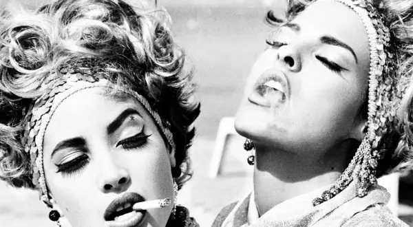 Iconica **Helmut Newton moda**: due donne con acconciature voluminose e make-up audace fumano sigarette, un'immagine di stile e provocazione.