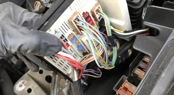 Ręka w rękawiczce manipuluje skrzynką z bezpiecznikami w Renault Scenic 2. Widać kolorowe przewody i oznaczenia bezpieczników.