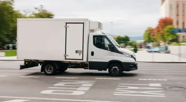 Biała chłodnia na podwoziu Iveco Daily, idealna do transportu wymagającego odpowiedniego prawa jazdy C1, jedzie po miejskiej ulicy.