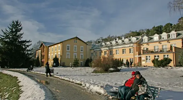 Żółty budynek sanatorium otoczony śniegiem, ludzie siedzą na ławce.