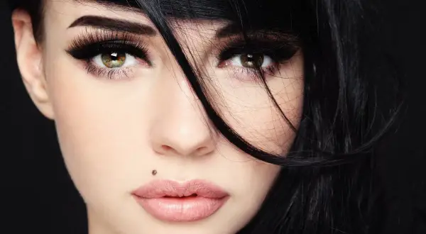 Kobieta z czarnymi włosami i makijażem podkreślającym oczy. Widoczny monroe piercing nad górną wargą.