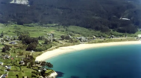 Vista aérea de la playa de Area, Viveiro, con casas dispersas entre verdes colinas y el mar turquesa.