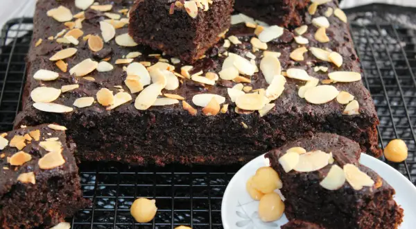 Pyszne brownie z ciecierzycy, posypane płatkami migdałów, wygląda apetycznie na kratce. Kilka kawałków podano na talerzyku z dodatkiem orzechów.