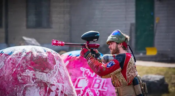 Gracz w stroju bojowym celuje z markerem, gotowy do akcji w laserowym paintballu. Za nim różowe dmuchane przeszkody.