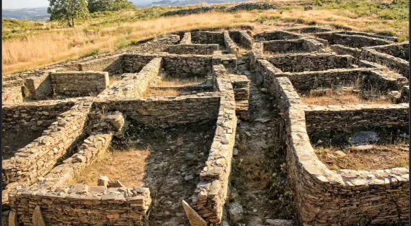 Ruinas de piedra del castro de Castromaior, con muros bajos y patios interiores, bajo un cielo azul y vegetación seca.