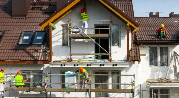 Pracownicy na rusztowaniach prowadzą termomodernizację budynku, ocieplając jego fasadę.