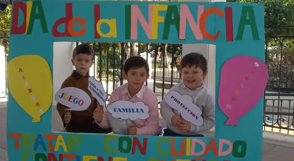 Niños sonríen tras un marco decorado para el Día de la Infancia, sosteniendo carteles con palabras como "Juego", "Familia" y "Protección".