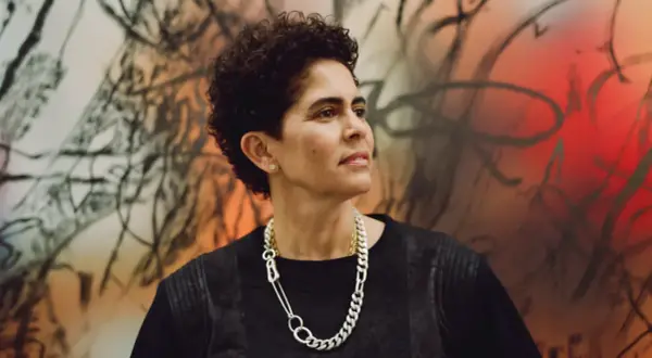 A striking portrait of artist Julie Mehretu, her gaze thoughtful, set against a vibrant, abstract background that echoes her dynamic style.
