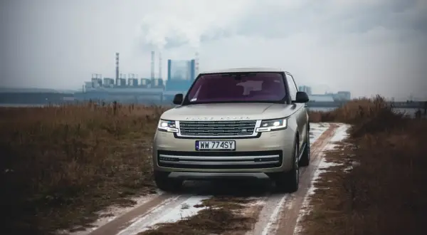 Złoty Range Rover Autobiography na błotnistej drodze, w tle przemysłowy krajobraz z kominami fabrycznymi.