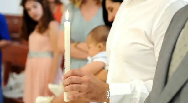 Un homme tient une bougie allumée, symbole de la lumière divine, lors d'un baptême. Il porte un bébé, prêt à accepter d'être parrain.