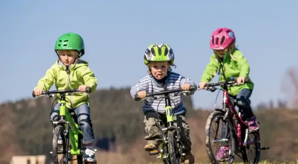 Trójka dzieci w kaskach uczy się jeździć na rowerze po trawiastym wzgórzu. Radość i pierwsze sukcesy w nauce jazdy na rowerze.