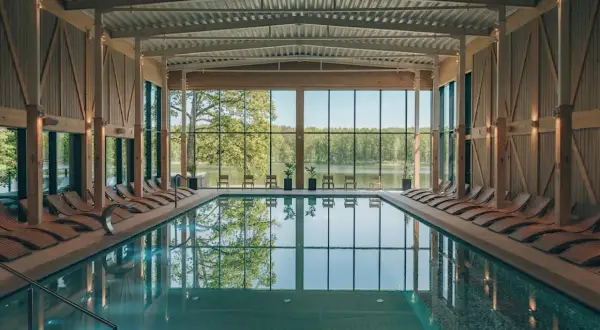 Basen w nowoczesnym spa z widokiem na jezioro i las. Drewniane leżaki i przeszklone ściany tworzą relaksującą atmosferę, idealną dla najlepszego spa w Polsce.