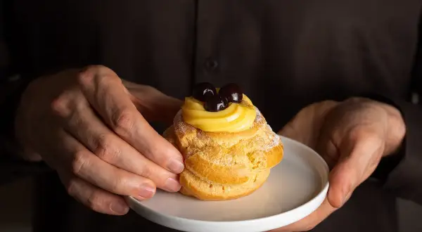 Mani che offrono una zeppola di San Giuseppe, un dolce con una ricca storia, guarnita con crema pasticcera e amarene.