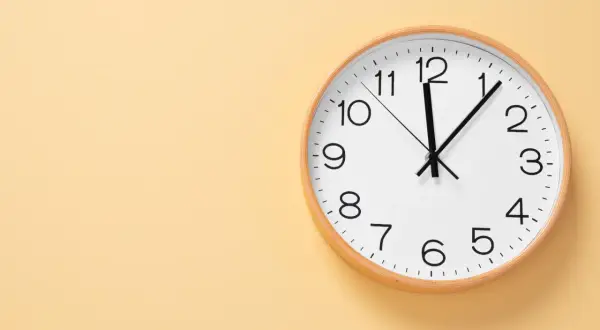 Reloj de pared blanco con marco de madera. Las manecillas marcan las 12:05. Ideal para aprender las horas en inglés, ejemplos.
