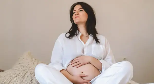 Kobieta w białej koszuli i spodniach siedzi na łóżku, obejmując brzuch. Jej zamyślona mina może sugerować tokofobia objawy.