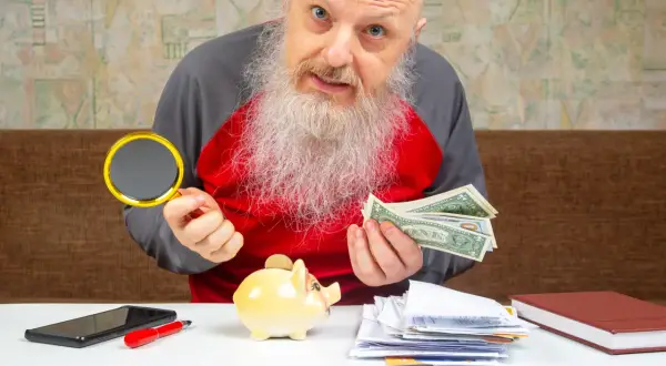 Starszy mężczyzna z lupą i dolarami zastanawia się, gdzie wymienić stare funty. Obok świnka-skarbonka i stos dokumentów.