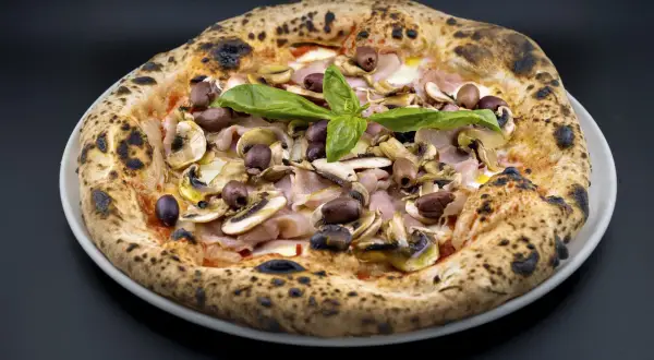 Pizza Capricciosa con champiñones, jamón y aceitunas negras, coronada con albahaca fresca. ¡Un verdadero capricho!