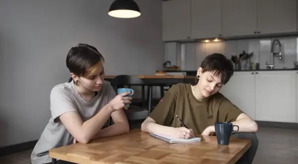 Dwie osoby przy stole, jedna pisze, druga pije kawę. Rozmowa o tym, jak załatwić zaświadczenie o dysleksji, może być wsparciem w nauce.
