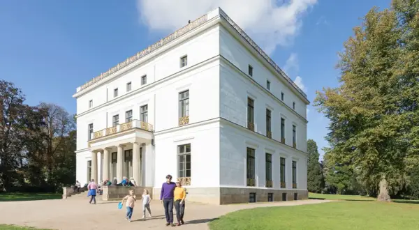 Familie spaziert durch den Jenisch Park vor einem weißen Herrenhaus mit Säulen und goldenen Verzierungen.