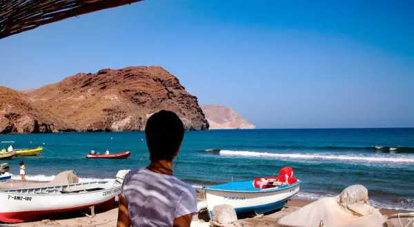 Persona contempla la **playa Las Negras** con barcas en la arena y el mar azul. Montañas áridas al fondo.