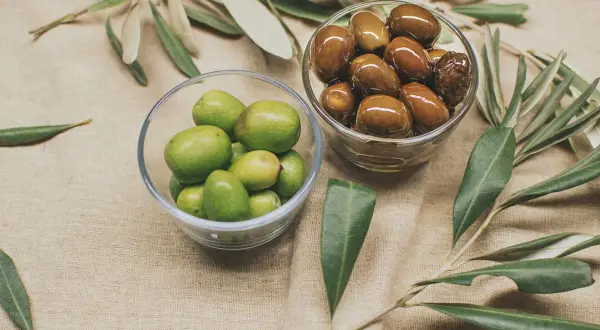 Due ciotole di vetro mostrano due tipi di olive: verdi e marroni, con rami d'ulivo sullo sfondo.